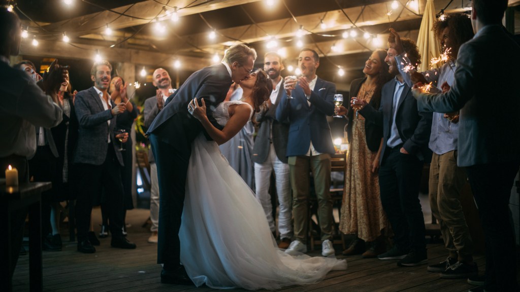 A bride and groom kiss on the dancefloor at their evening wedding reception surrounded by celebrating friends and string lights.
