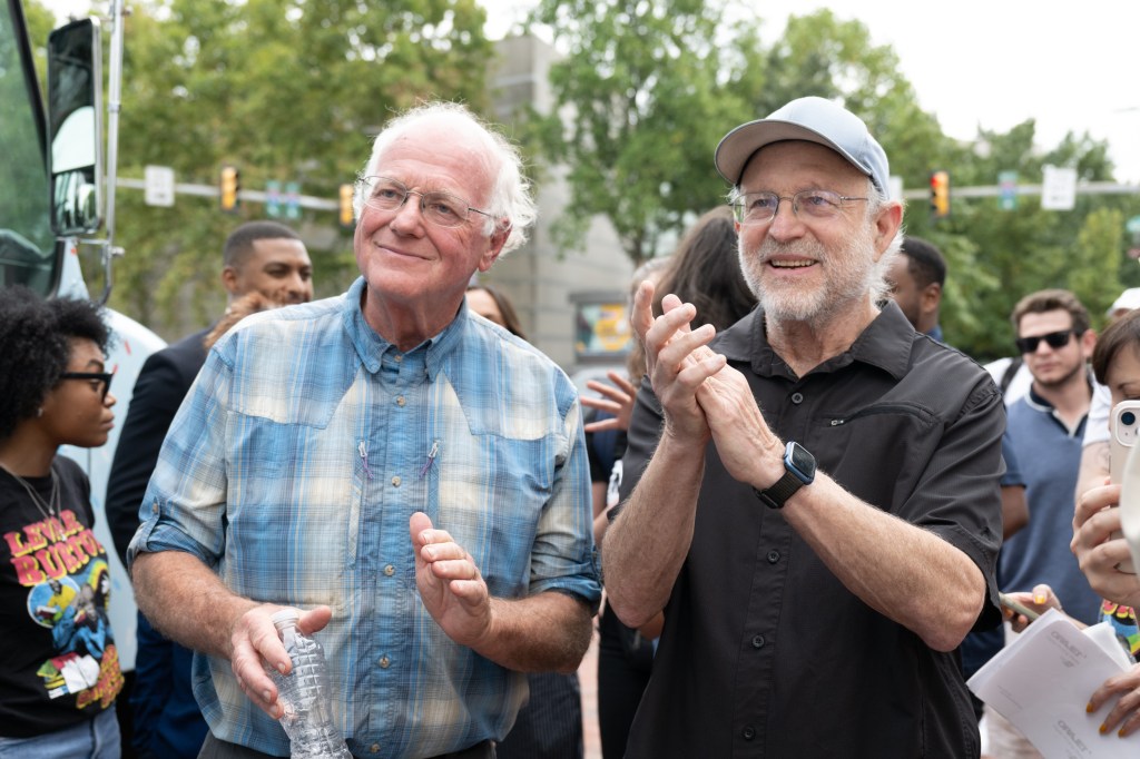 Ben Cohen and Jerry Greenfield clapping while holding a water bottle and wearing a hat respectively.