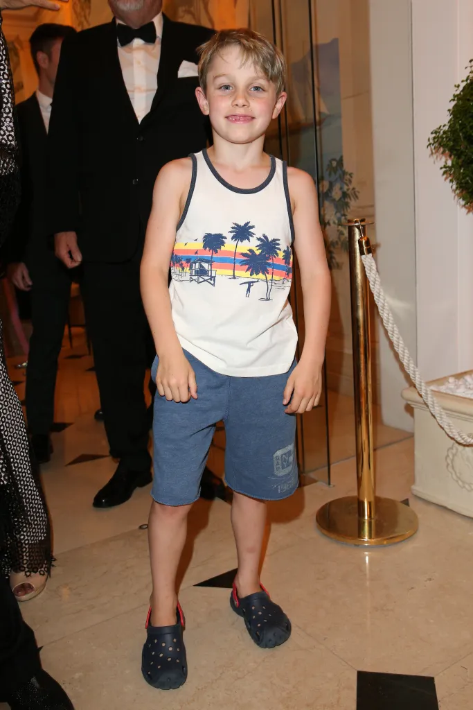 Benjamin Travolta, son of John Travolta, wearing a tank top with a beach scene, blue shorts, and dark blue Crocs with red straps.