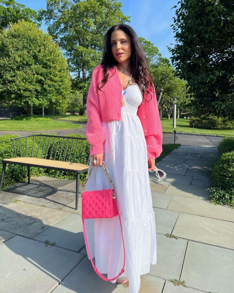 Bethenny Frankel standing outdoors wearing a white dress and a pink cardigan, holding a pink Louis Vuitton bag.