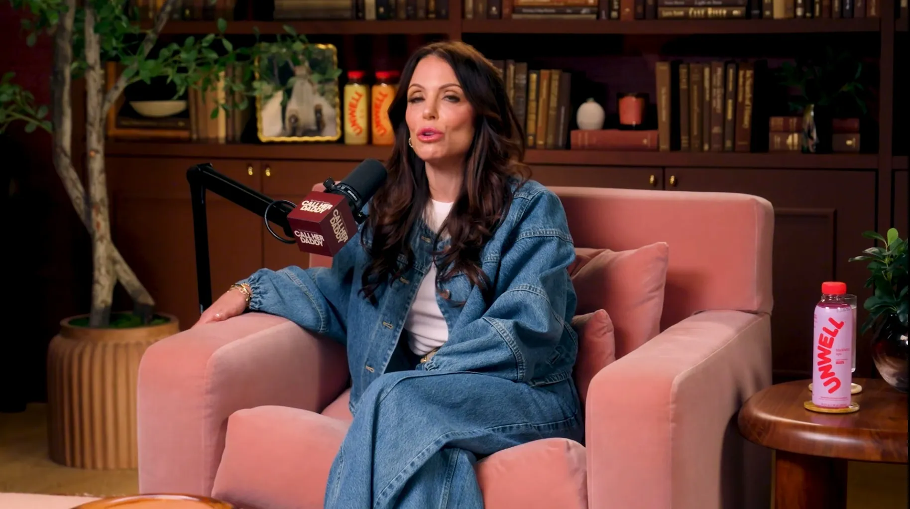 Bethenny Frankel in a denim outfit sitting in a pink armchair, speaking into a 