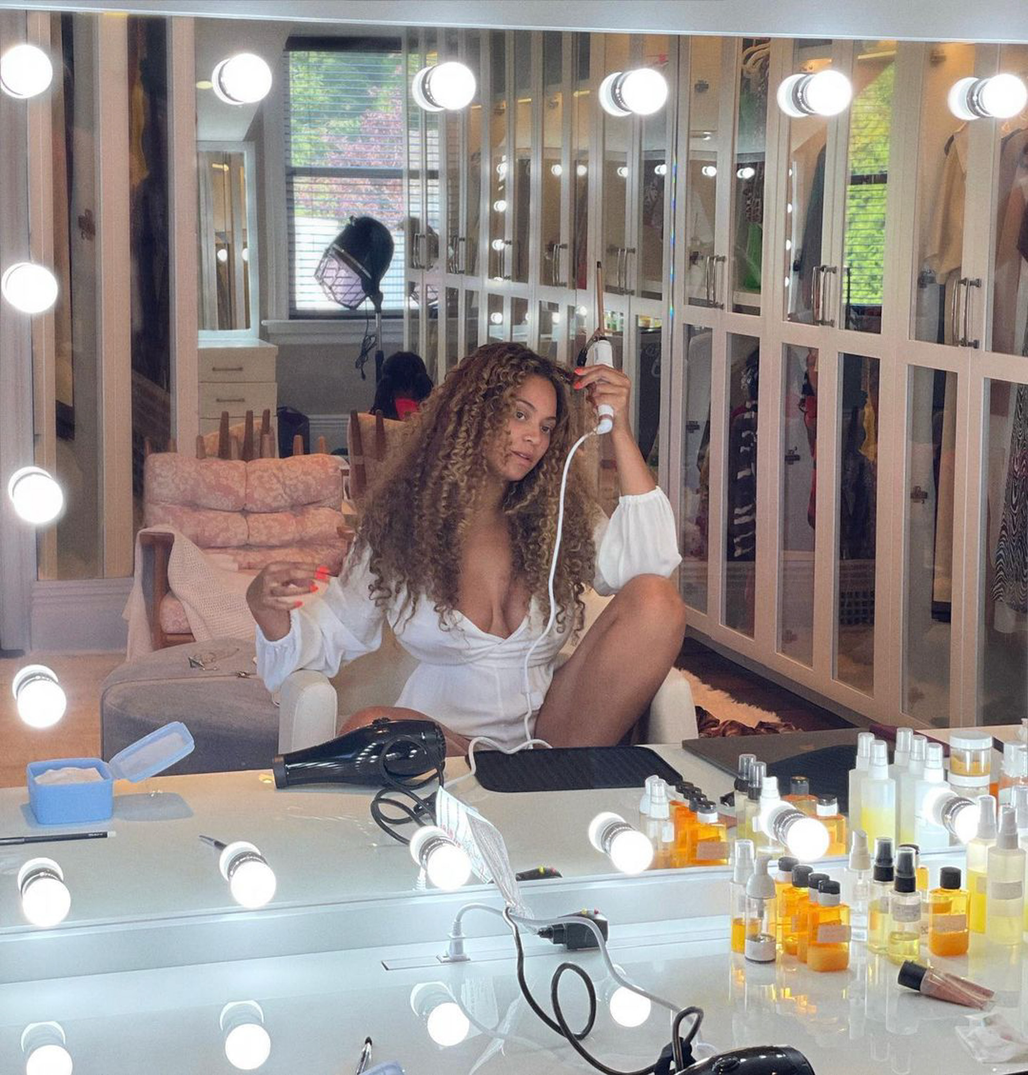 Beyoncé doing her hair with a curling iron in a mirror surrounded by hair products.