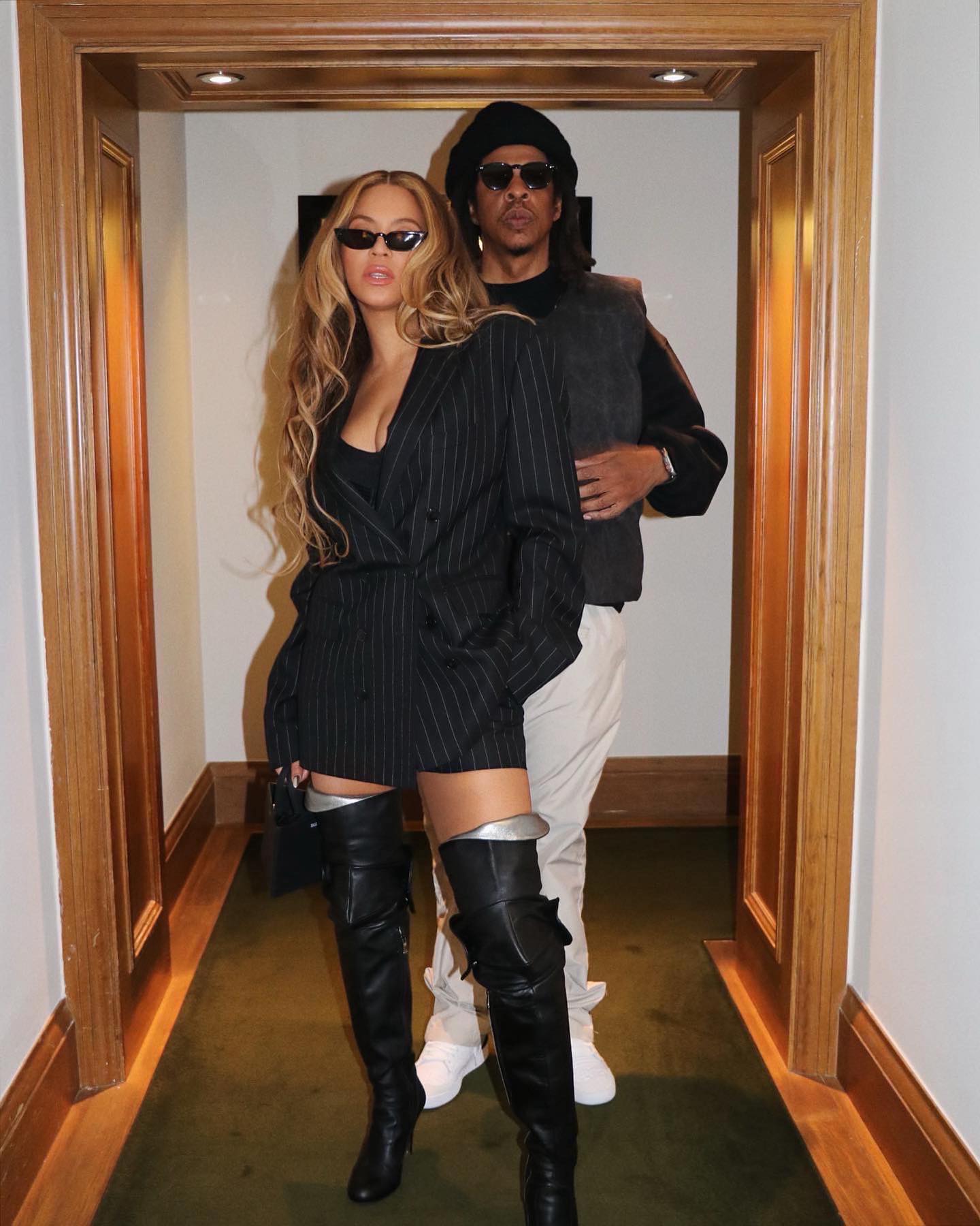 Beyoncé in a pinstripe blazer and thigh-high boots, with Jay-Z behind her in a black hat and vest.