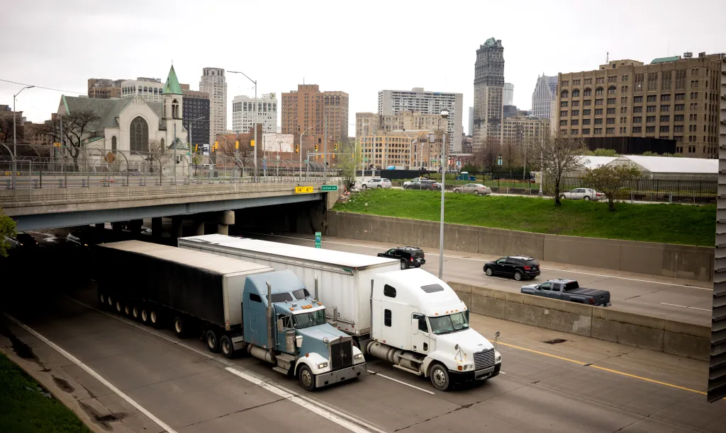Two semi-trucks on an Interstate 75 highway with Detroit's city skyline in the background.