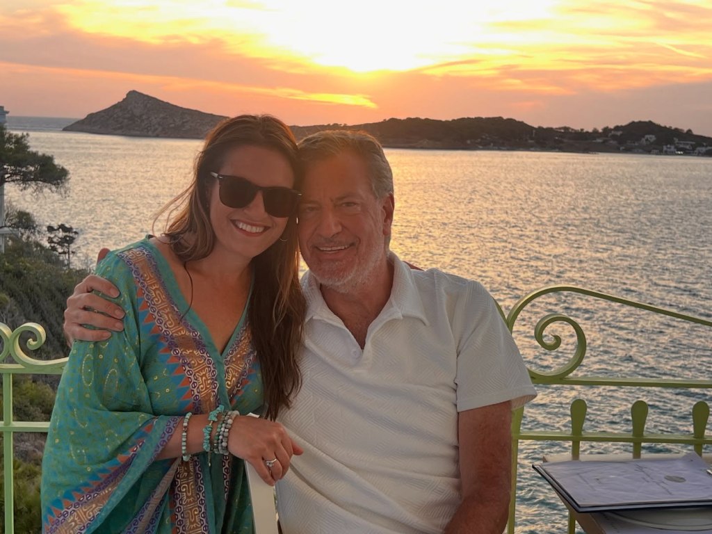 Bill de Blasio and Nomiki Konst posing together with a sunset over the water in the background.