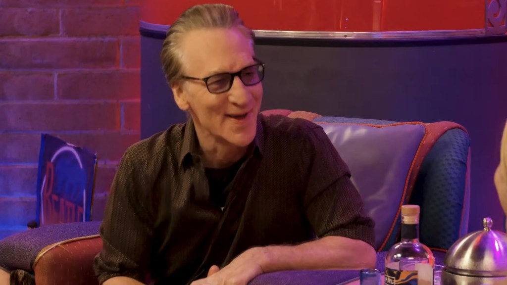 Bill Maher laughing while sitting in a blue and orange chair next to a bottle of alcohol.