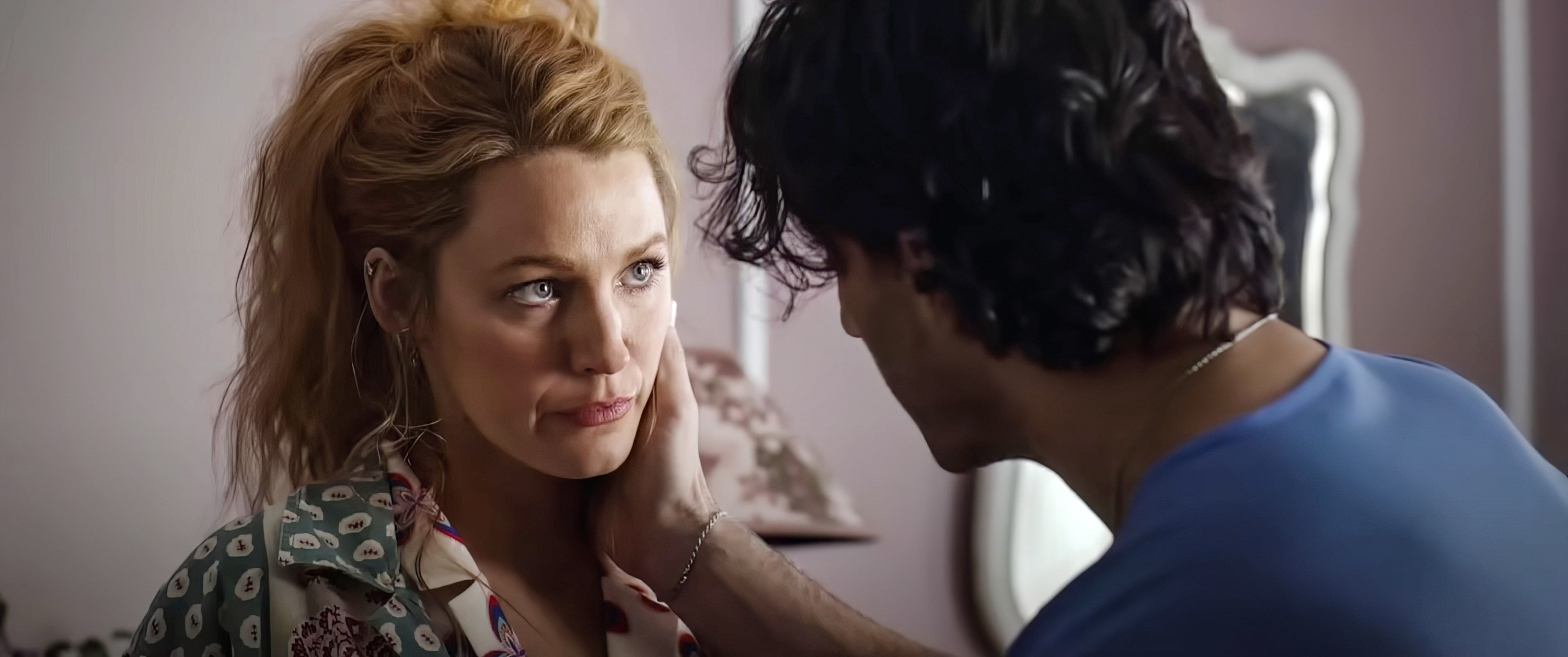 Blake Lively with her hair in a messy bun, wearing a patterned blouse, is held by her chin by Justin Baldoni in a blue shirt.