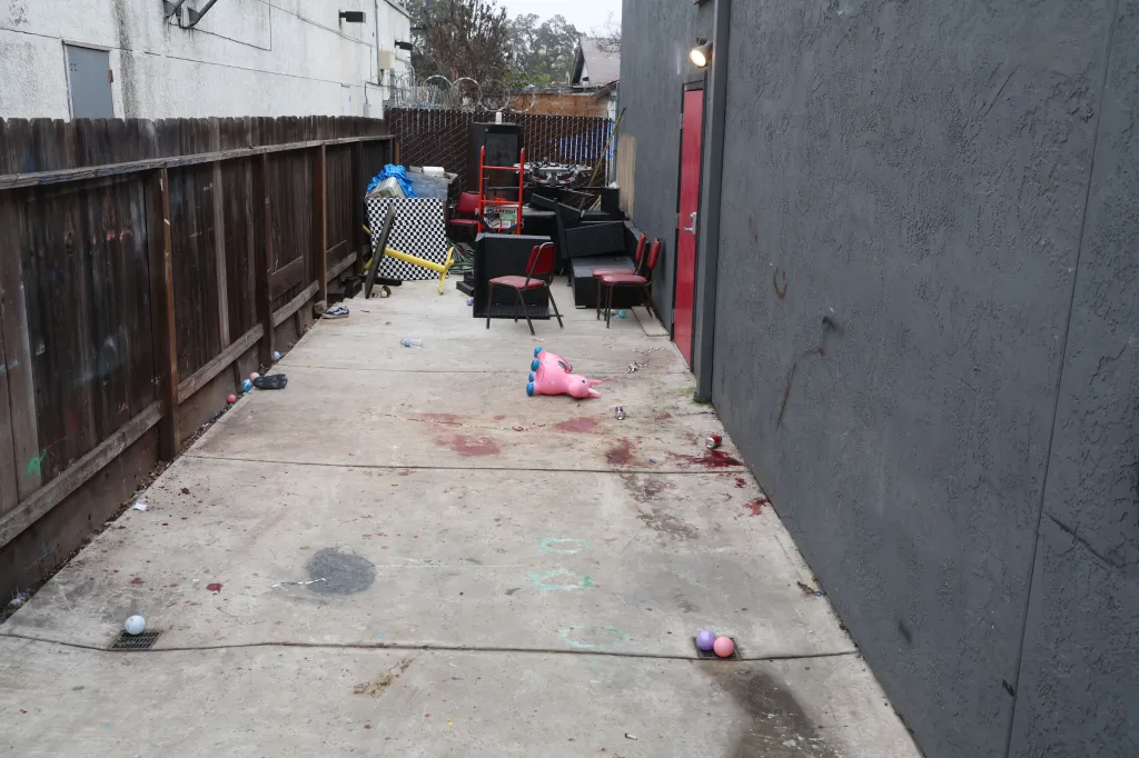 Back exit area of a building showing a wooden fence, debris, chairs, a bouncy toy, and blood stains on the concrete floor.