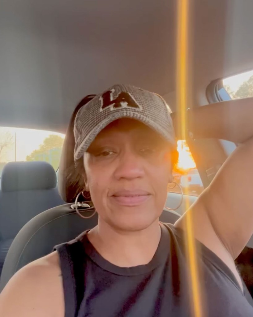 A woman in a car wearing a hat and black shirt, smiling, with sun shining through the window.