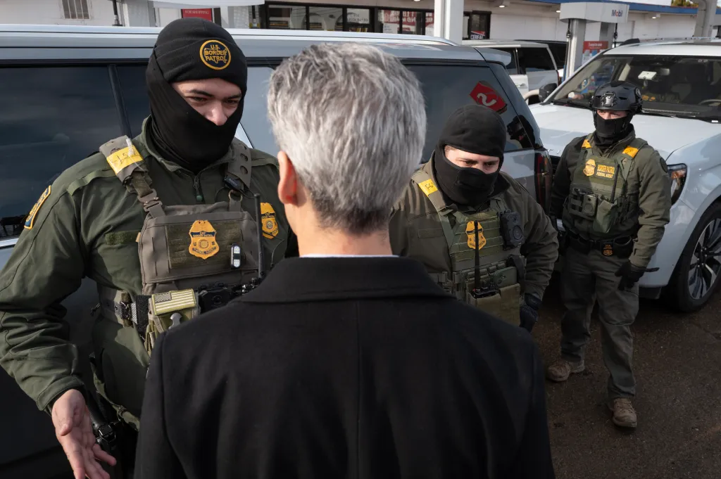 Three Border Patrol agents, dressed in tactical gear and balaclavas, confront Evanston Mayor Daniel Biss at a gas station.