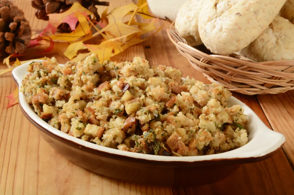 A bowl of turkey dressing next to a basket of multigrain rolls, pine cones, and autumn leaves on a wooden table.
