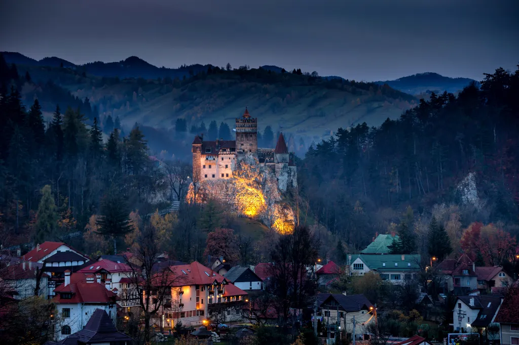 Bran Castle, known as Dracula's Castle, illuminated at twilight, with a village in the foreground and forested mountains behind, in Transylvania, Romania.