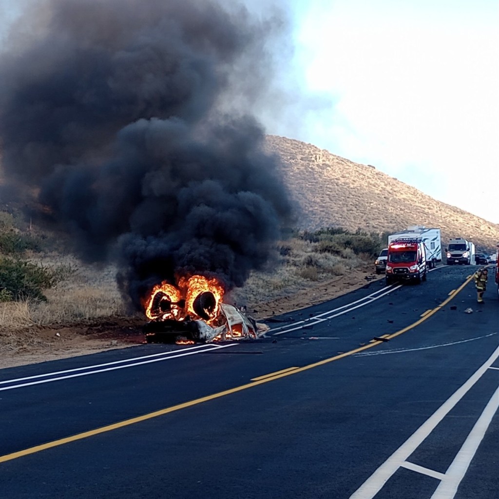 A car on fire with thick black smoke on the side of a highway, with an ambulance and other vehicles in the background.