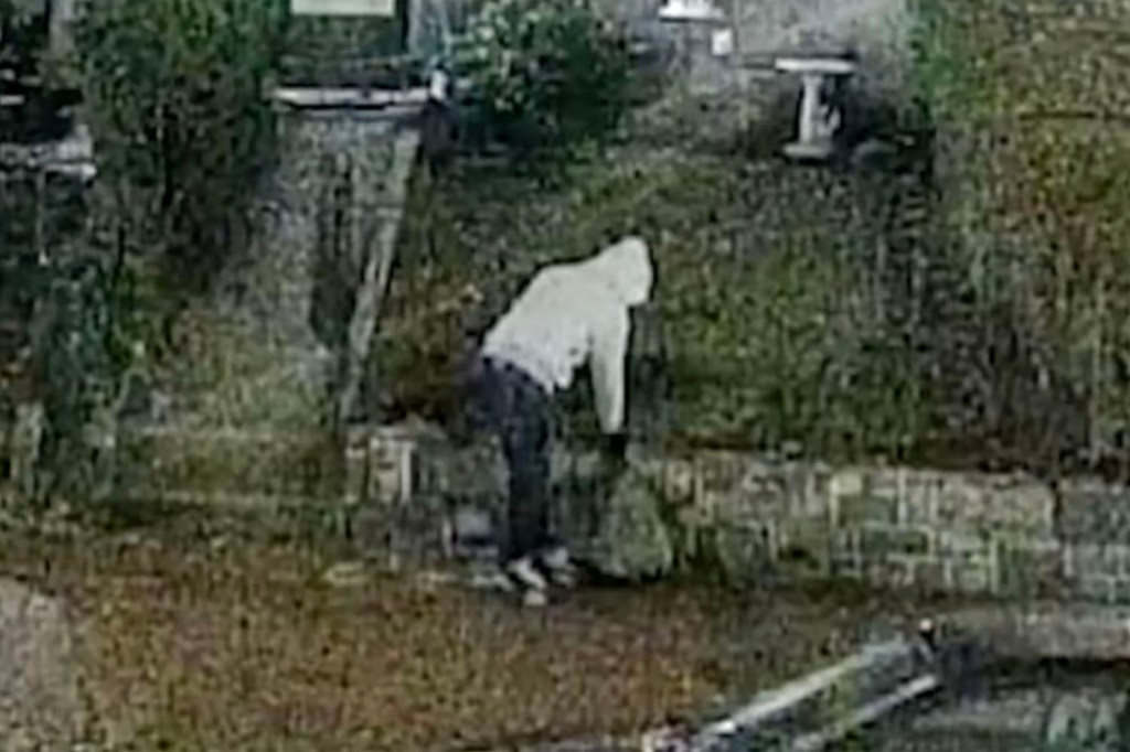 A still from FBI video footage showing a person in a light-colored hooded sweatshirt placing an object near a brick wall.