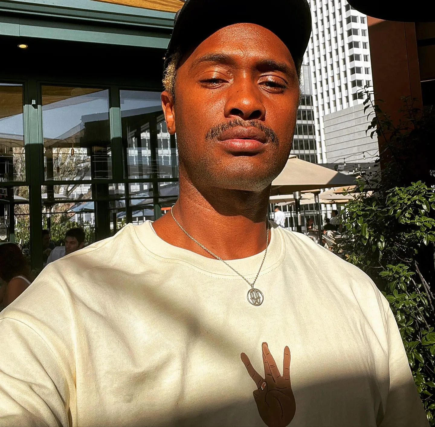 Brian McKnight Jr. looking into the camera wearing a black cap, a beige t-shirt with a peace sign, and a silver necklace.