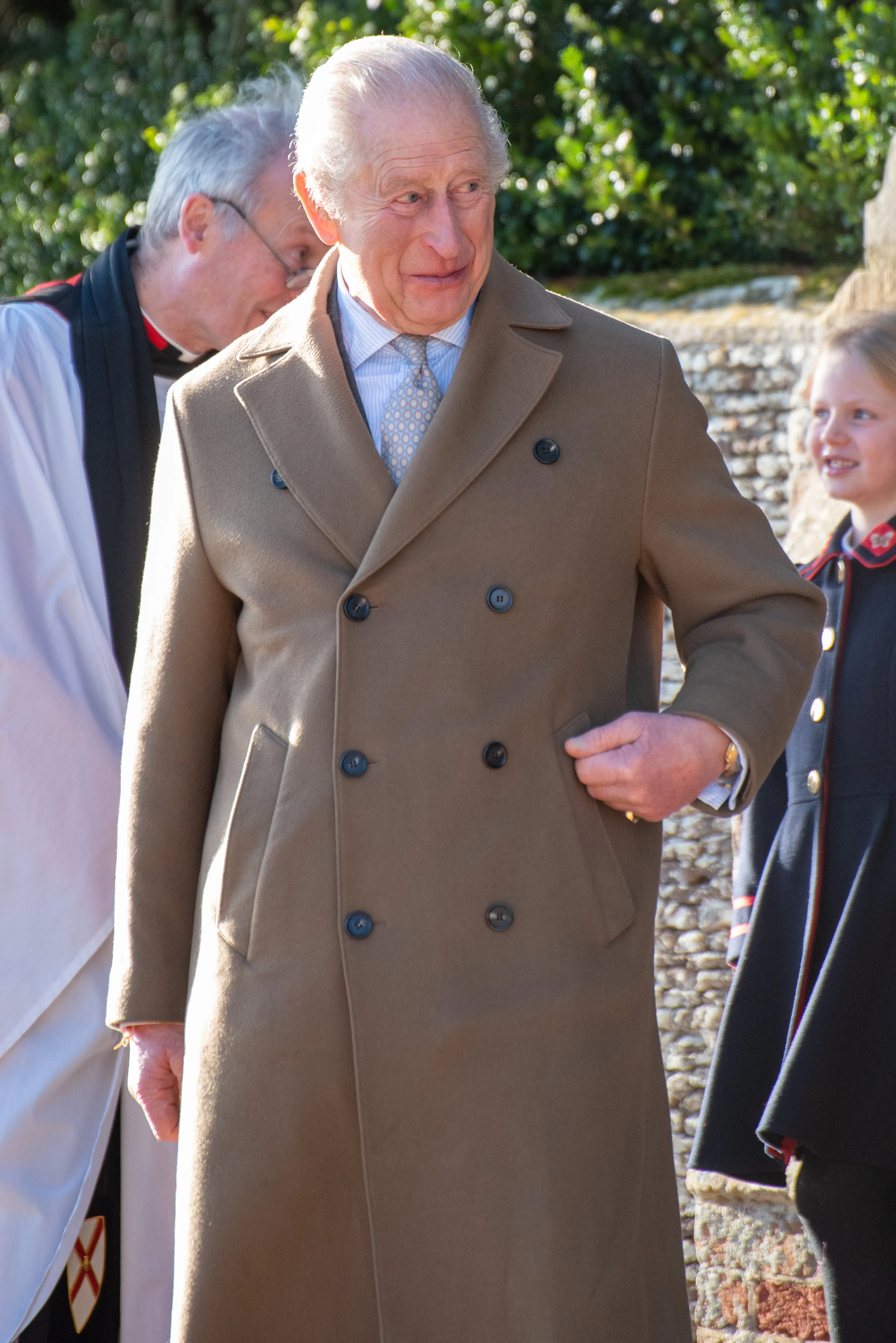 King Charles III in a tan overcoat with his hand in his pocket, looking to the right.