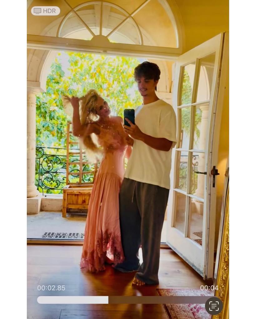 Britney Spears in a peach dress and Jayden Federline in a white t-shirt posing for a selfie indoors with an outdoor background.