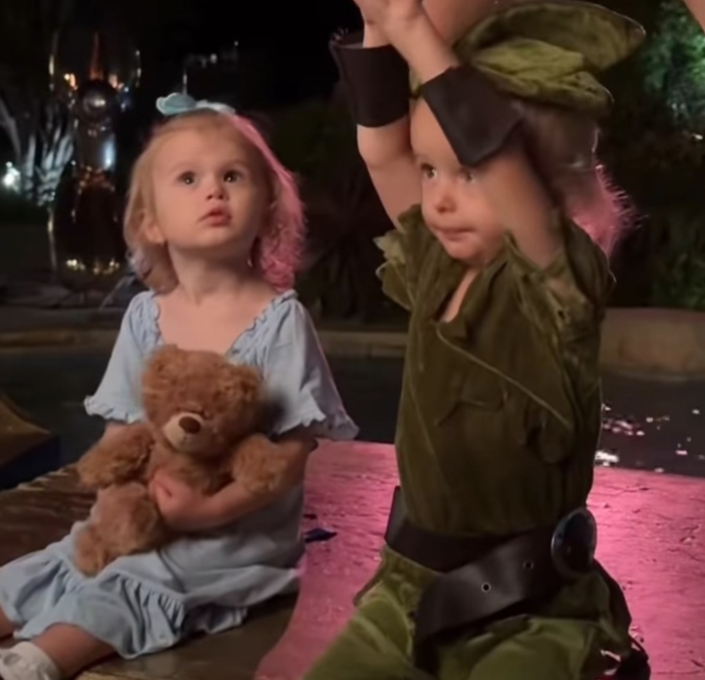 Two young girls in costumes, one in blue holding a teddy bear and the other in green.