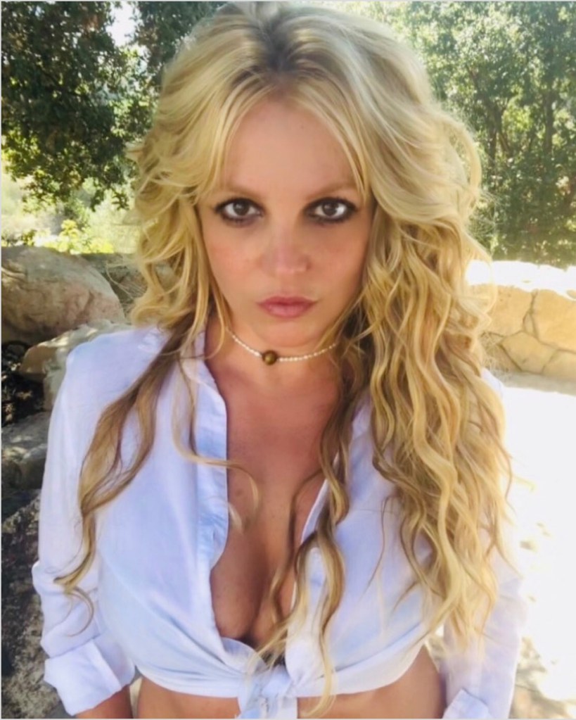 Britney Spears with long, wavy blonde hair, wearing a white unbuttoned shirt tied at the waist.