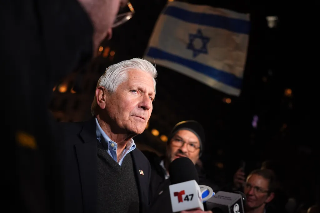 Bruce Blakeman speaks into microphones with an Israeli flag in the background.
