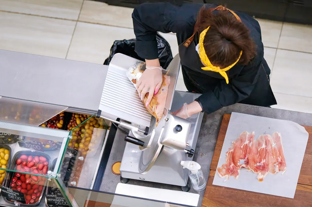A female deli worker in a black uniform and yellow headband slicing prosciutto with a meat slicer.
