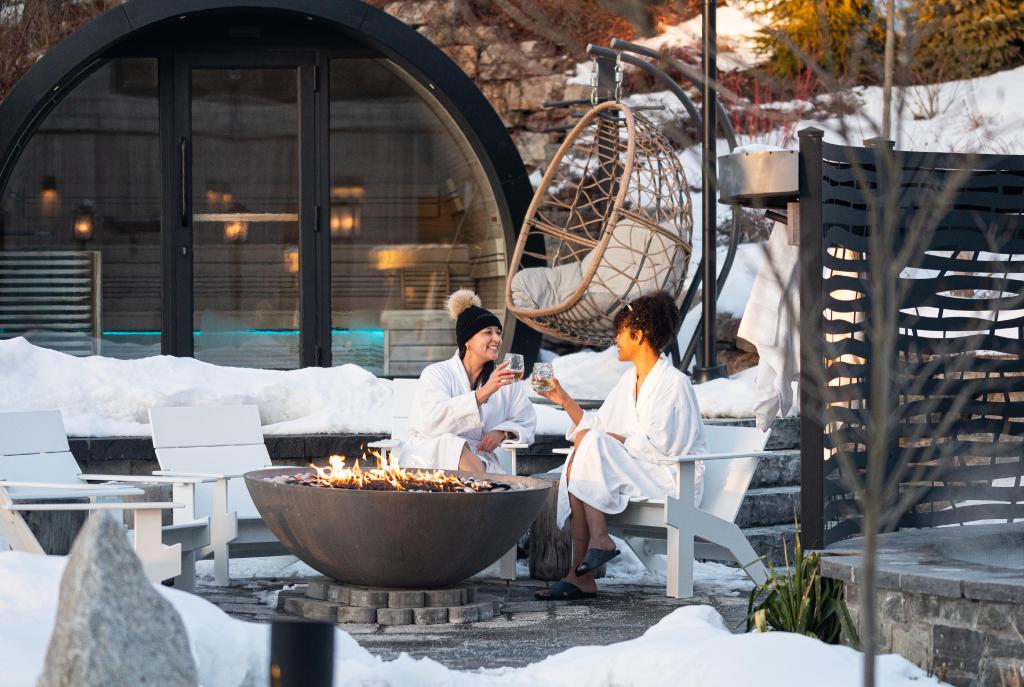 Two women in robes toast drinks by a fire pit at a snowy Nordic spa.