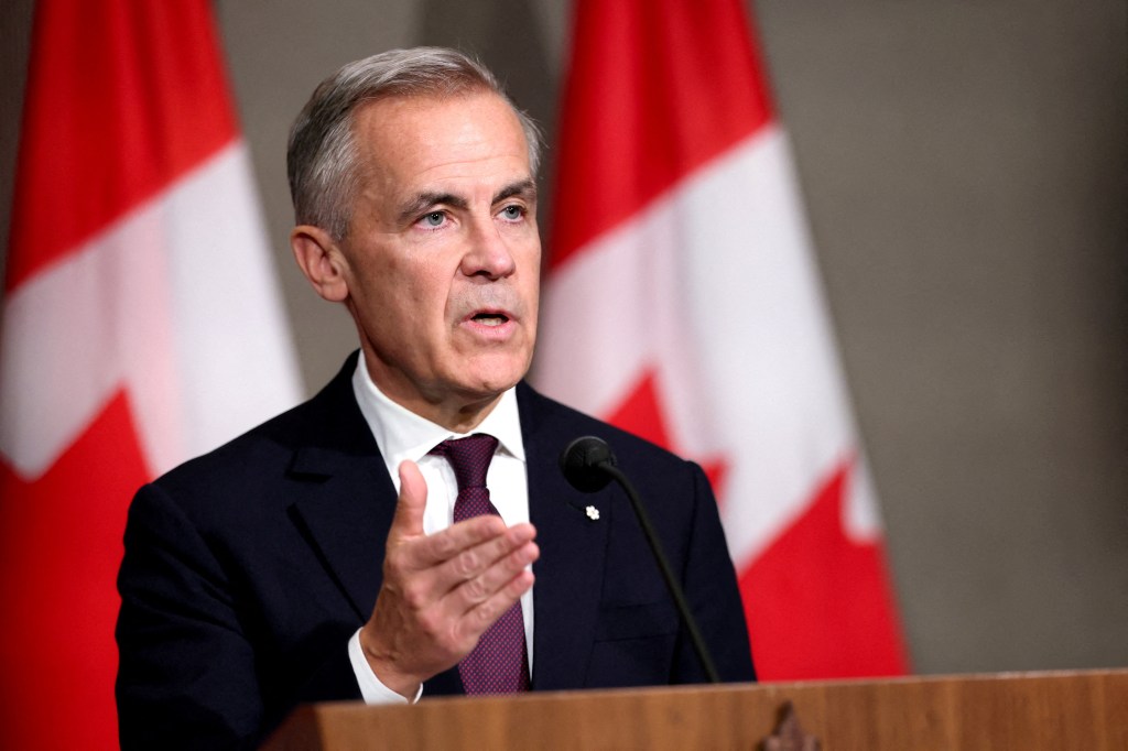 Canada's Prime Minister Mark Carney speaking at a press conference, with Canadian flags in the background.