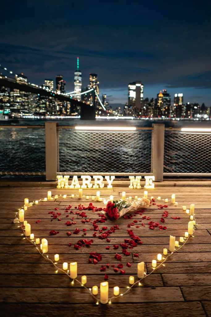A heart outlined by candles and string lights, with red rose petals and a bouquet of red roses inside, and illuminated 