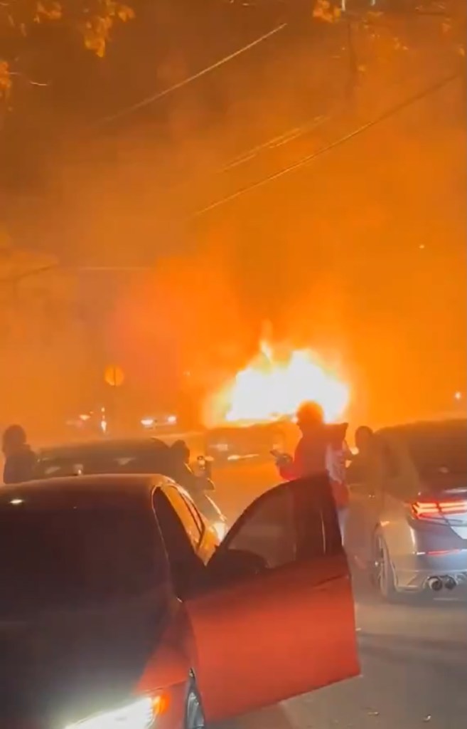 Car on fire in the street at night with a group of people standing around it.