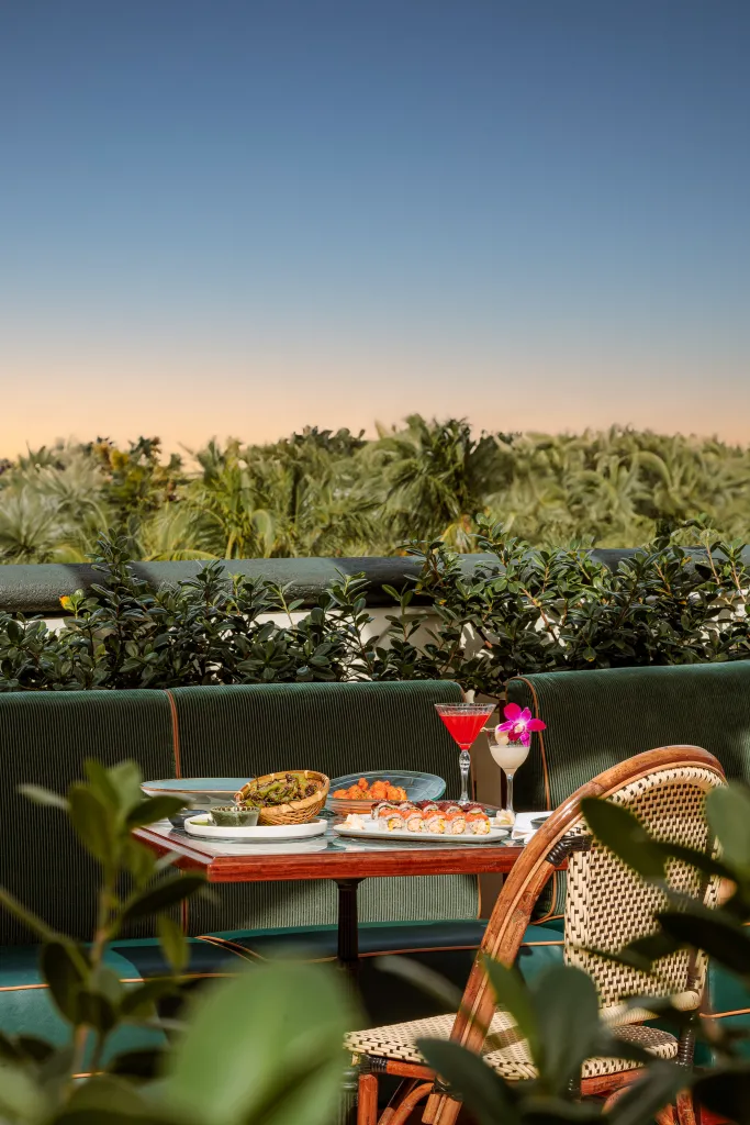 Outdoor dining table with sushi, cocktails, and other food, set against a backdrop of lush green foliage and an evening sky.