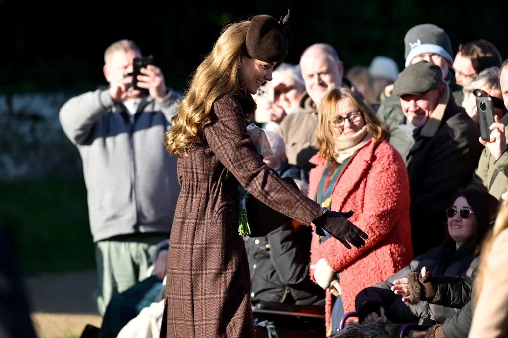 Catherine, Princess of Wales, in a plaid coat and brown hat, greets a crowd of people, some holding up phones to take pictures.