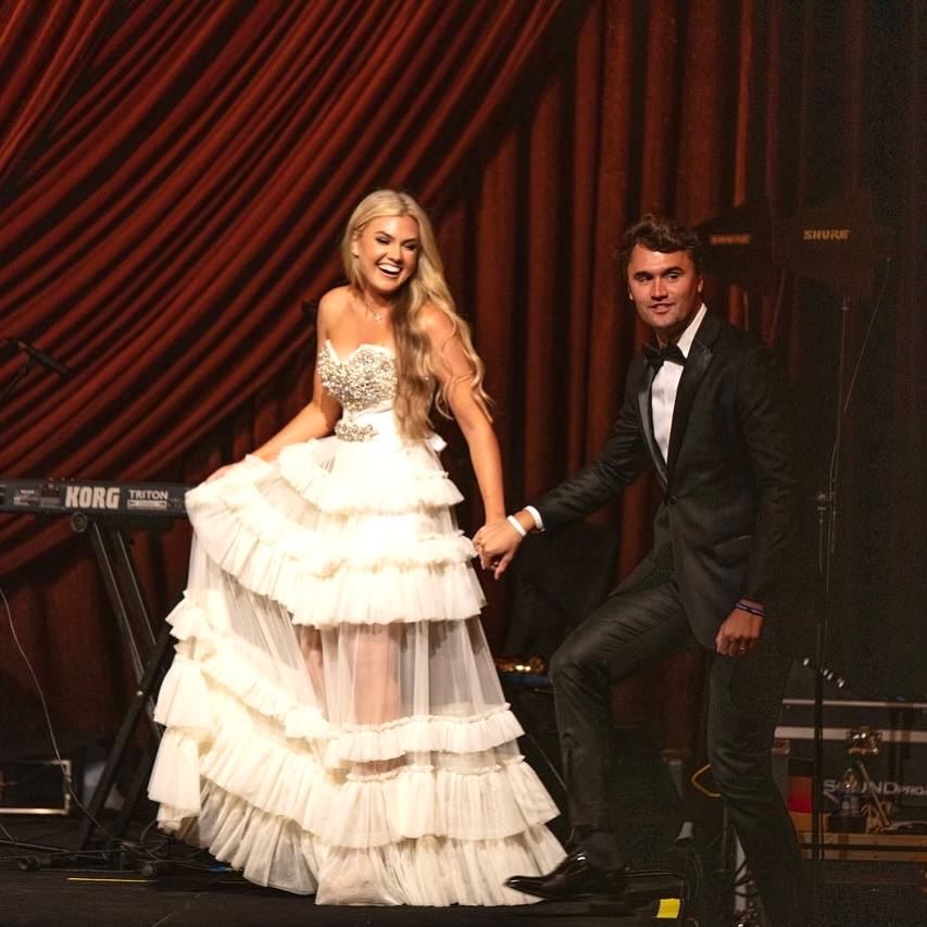 Erika Kirk in a white dress and Charlie Kirk in a black tuxedo, holding hands on a stage.