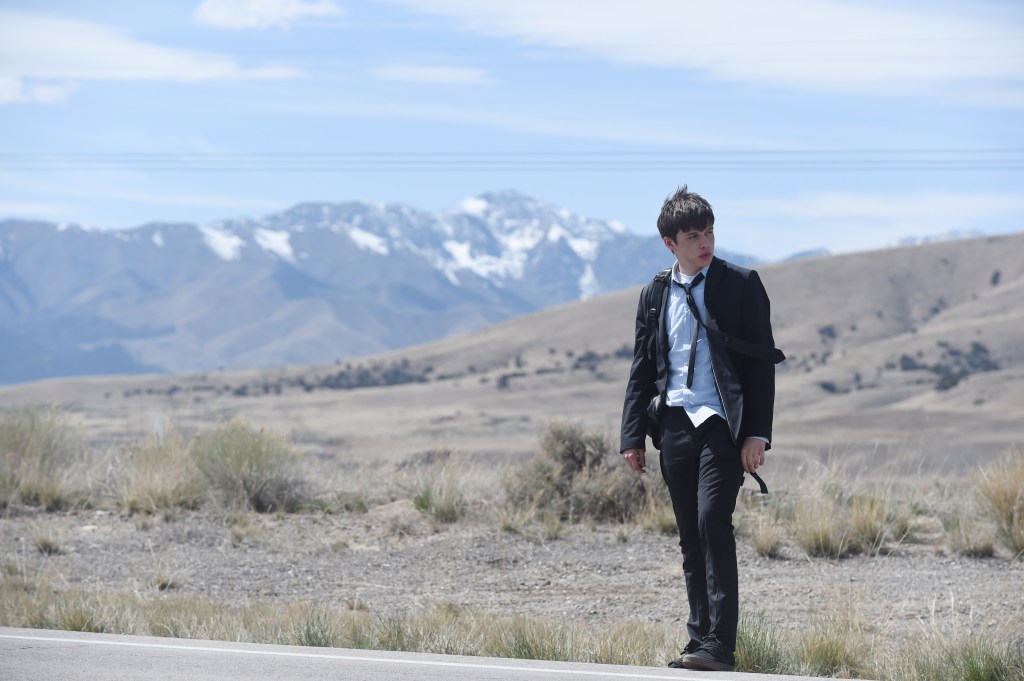Nick Robinson as Charlie in a black suit, light blue shirt, and backpack, standing on a road with snowy mountains in the background.