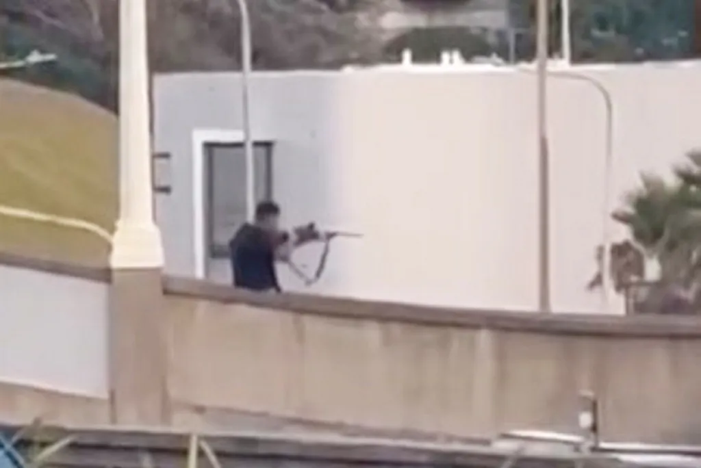 A man in a dark shirt shooting a rifle from a bridge.
