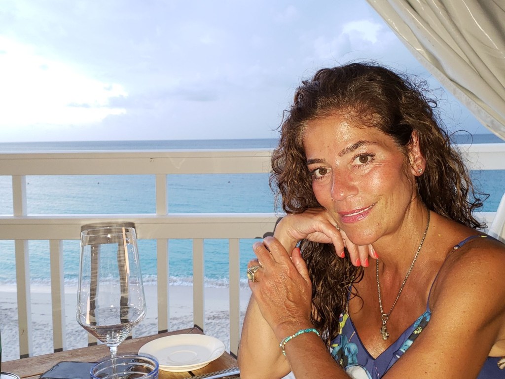 A woman with curly brown hair in a blue dress leans on a table with a white wine glass and a plate, with a beach and ocean in the background.