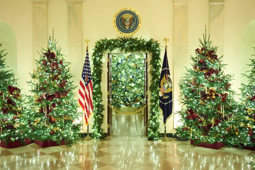 Christmas trees decorated with lights, red and gold ornaments, and candles fill the Cross Hall of the White House, with American and Presidential flags flanking a doorway.