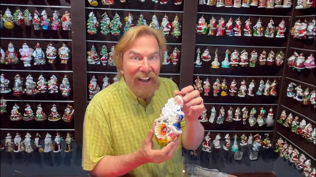 Christopher Radko holding a large Christmas ornament in front of shelves filled with smaller Christmas ornaments.