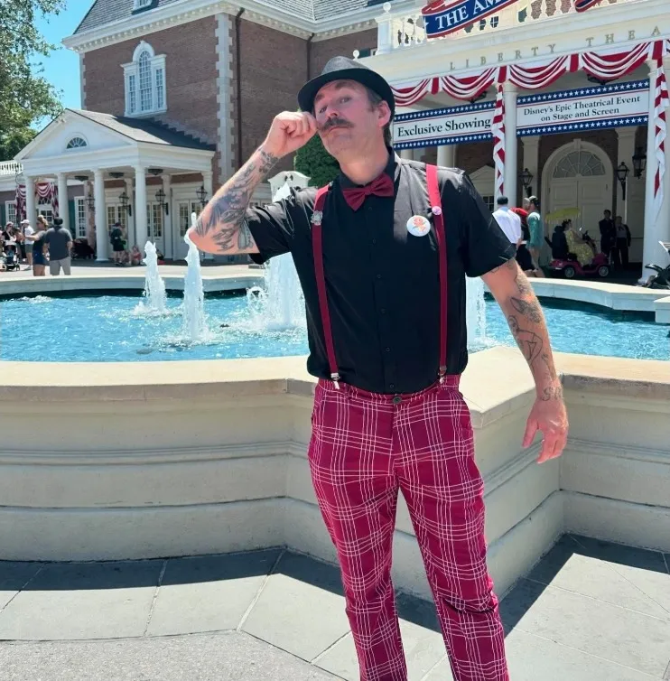 Man posing in a top hat, black shirt, red suspenders, and red plaid pants in front of a fountain and 