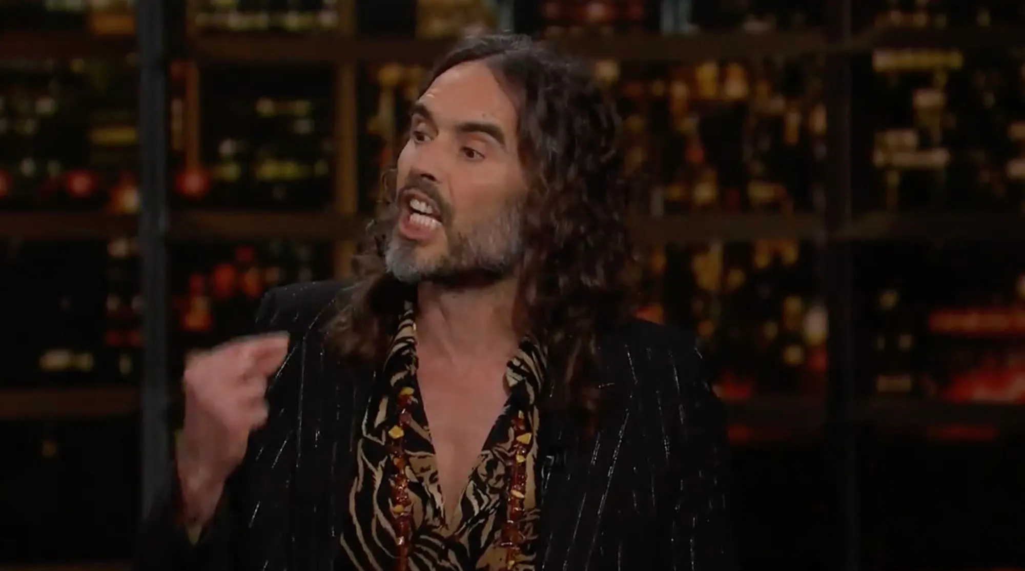 Russell Brand speaking at a show, wearing a patterned shirt and necklaces.