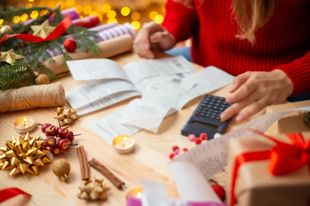A woman calculating Christmas expenses using receipts and a calculator, with holiday decorations around.