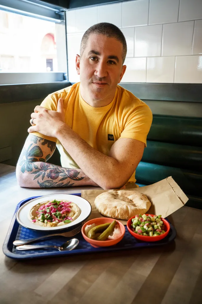 Michael Solomonov sitting at a table with a tray of hummus, pita, and side dishes.
