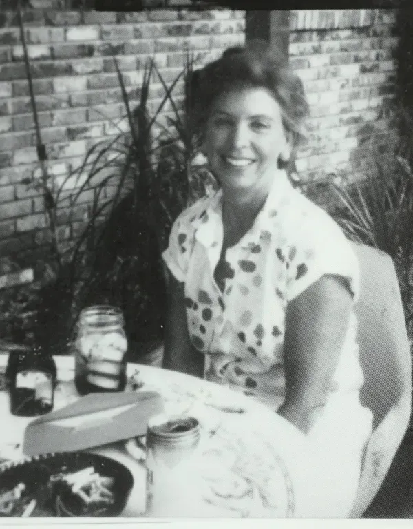 A smiling woman in a polka-dotted shirt sits at an outdoor table with jars.