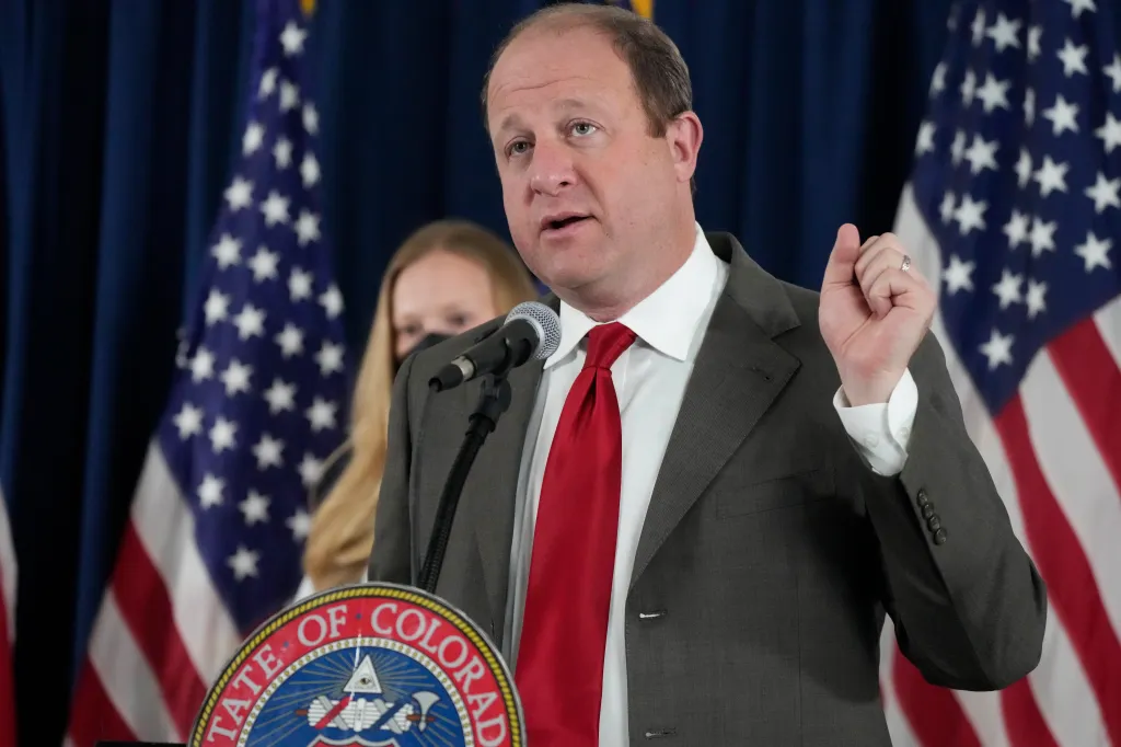 Colorado Gov. Jared Polis speaking at a news conference with state epidemiologist Rachel Herlihy listening in the background.