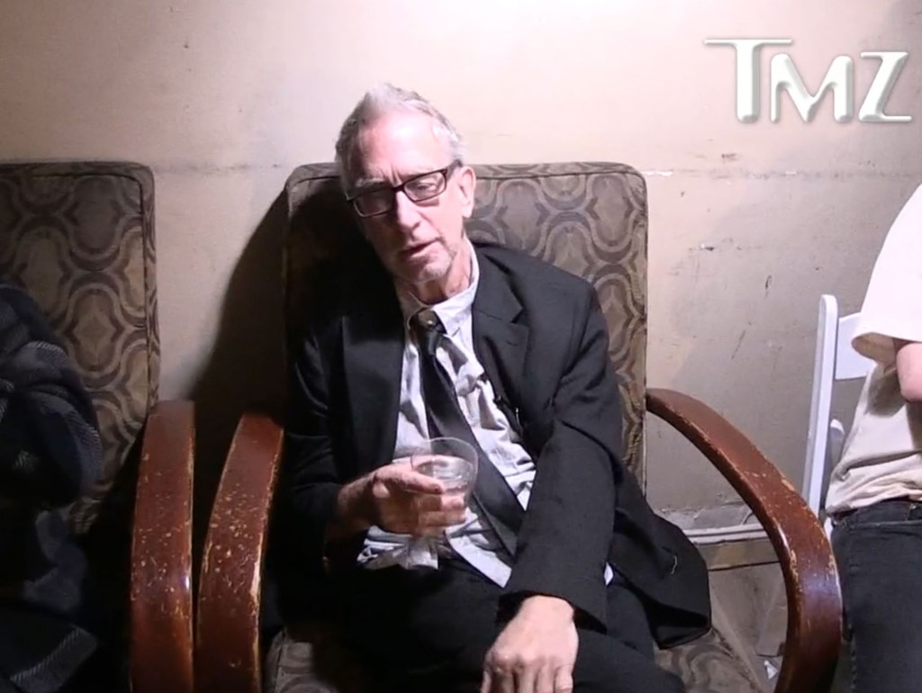 Comedian Andy Dick seated in a patterned armchair, holding a glass.