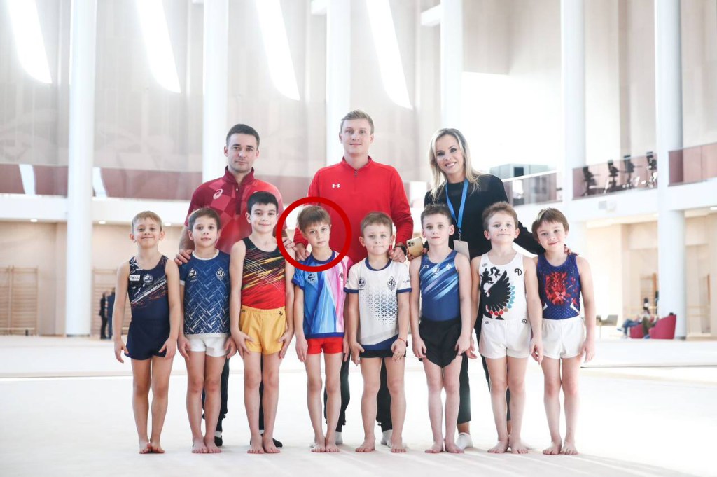 Group photo of eight young gymnasts with three adults in a large indoor space.