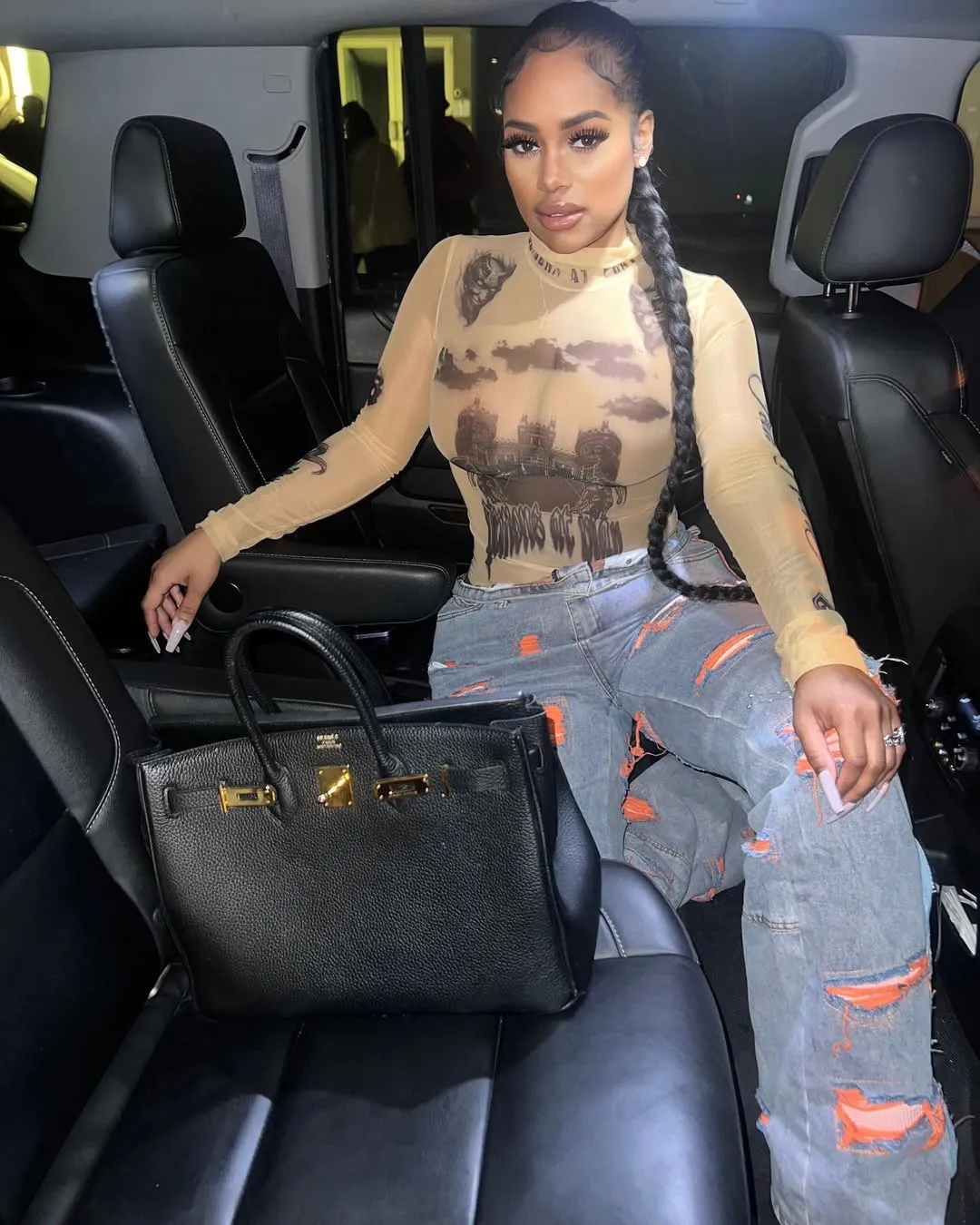 Joanna Donnejour with a long braided ponytail sits in the back seat of a car, wearing a sheer long-sleeved top with a landscape design, ripped jeans, and with a black handbag beside her.