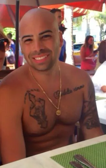 Gene Borello, bald and shirtless, with tattoos on his chest and shoulder, wearing a gold chain and medallion, smiling.