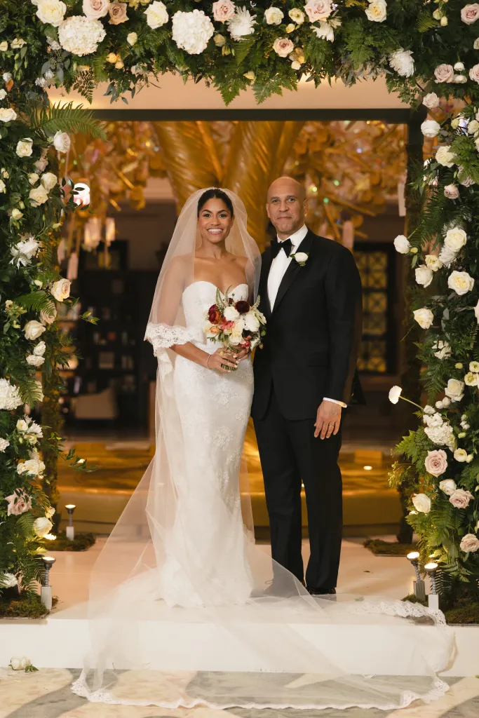 Cory Booker and Alexis Lewis pose together in wedding attire under a floral arch.