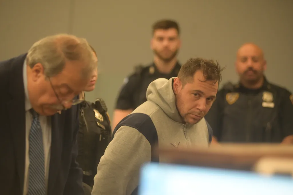 John Pilaccio, in a grey and blue hoodie, looks at the camera in a courtroom with other people out of focus in the background.