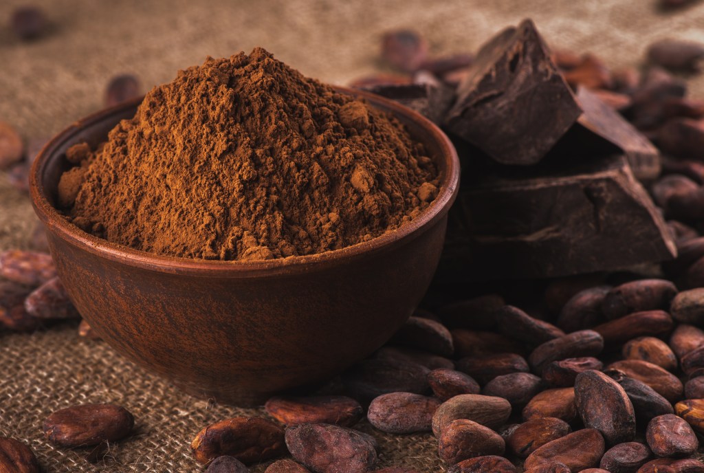 Cocoa powder in a brown ceramic bowl, raw cocoa beans and chocolate on burlap.